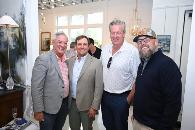 Four men smiling indoors, one wearing a grey blazer, another a tan blazer, and two in casual shirts.