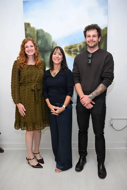 Three people stand in front of a landscape painting. A woman in a green dress, a woman in a navy