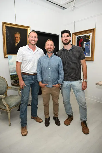 Three men smiling in a room with artwork on the walls and a vintage chair.