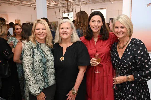 Four women smiling at an event. One wears a snakeskin print top, another a black dress, a third a
