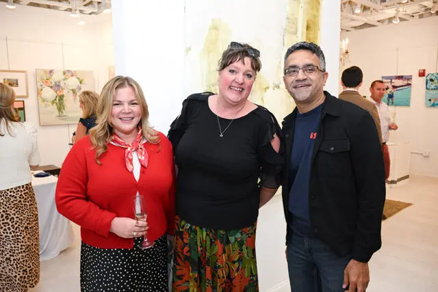 Three people smiling at an event. Woman in red cardigan, woman in black shirt, man in black jacket.