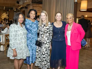 Five women posing for a photo at an event. The woman on the far right wears a bright pink suit.