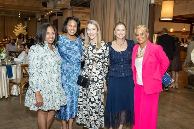 Five women posing for a photo at an event. The woman on the far right wears a bright pink suit.