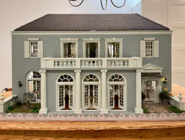 Gray dollhouse with white trim, shutters, balcony, and arched French doors.
