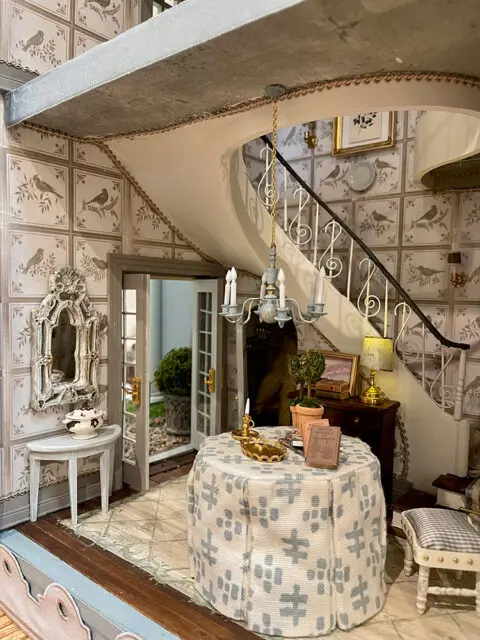 Dollhouse interior with bird-patterned wallpaper, a curved staircase, and a round table covered in