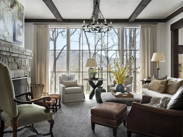 Living room with stone fireplace, large windows overlooking trees, and comfortable seating.