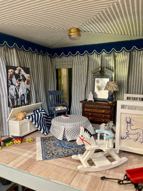 Dollhouse room with striped curtains, a rocking horse, toy chest, and patterned rug.