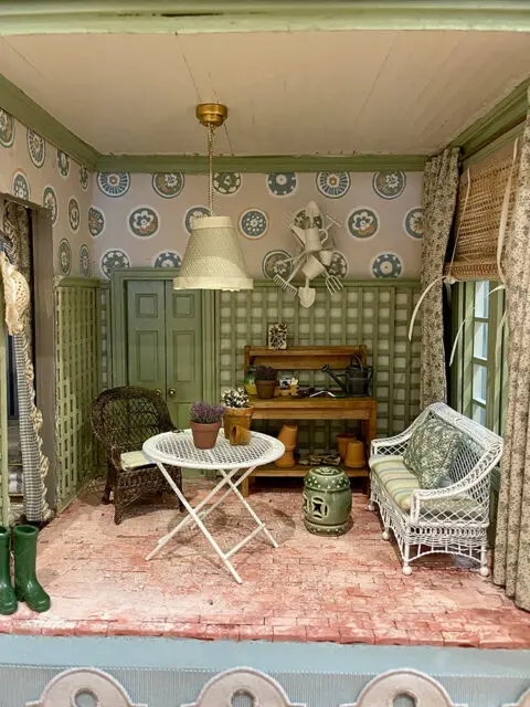 Dollhouse sunroom with wicker furniture, white table, gardening tools, and patterned wallpaper.
