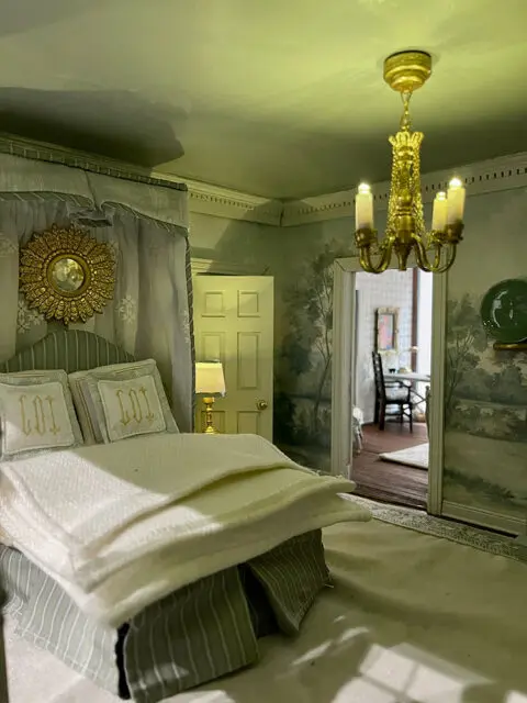 Dollhouse bedroom with four-poster bed, gold sunburst mirror, and mural wallpaper.