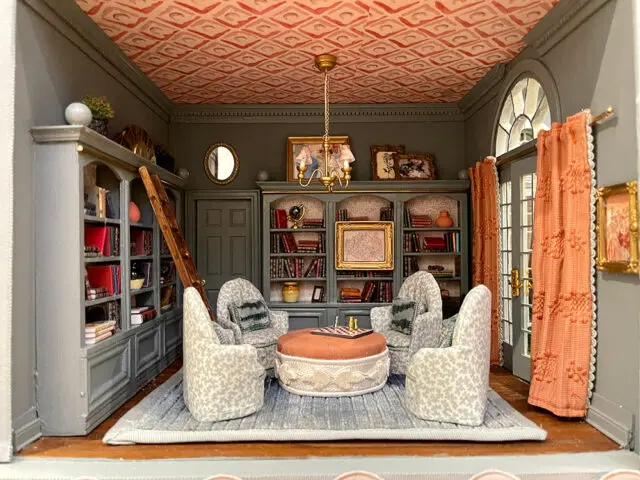 Miniature library with gray walls, built-in bookshelves, patterned ceiling, and upholstered chairs.