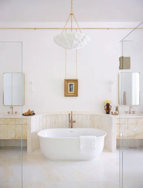 White freestanding bathtub with brass faucet, flanked by marble vanities and glass shower