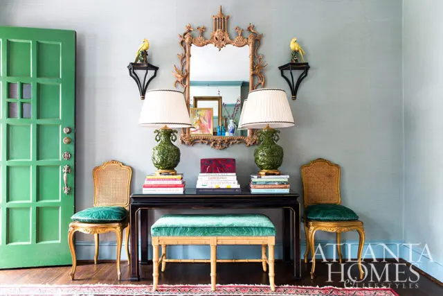 Green door, console table with green lamps, books, and chairs against blue grasscloth wallpaper.
