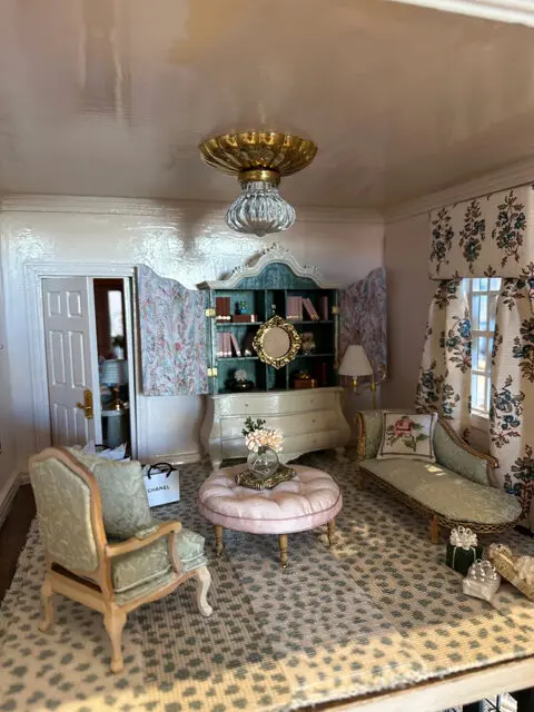 Dollhouse living room with green armchair, pink ottoman, chaise lounge, and patterned rug.