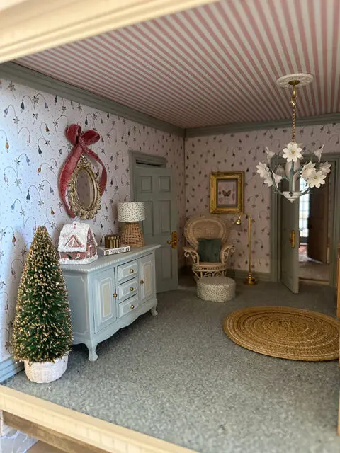 Dollhouse room with striped pink ceiling, floral wallpaper, blue dresser, and wicker chair.
