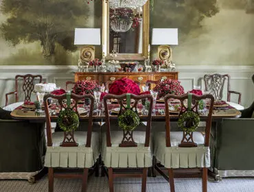 Dining room with a long table set for a holiday meal, featuring green velvet chairs and floral