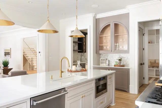 White kitchen island with brass faucet and microwave, gray arched cabinets with glass doors.