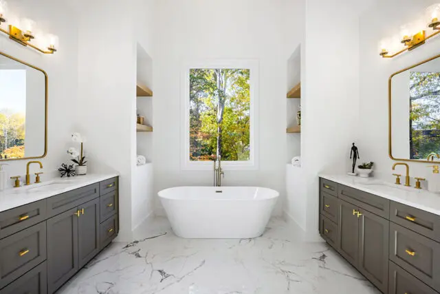 White freestanding bathtub with gold faucet, flanked by gray vanities with marble countertops and