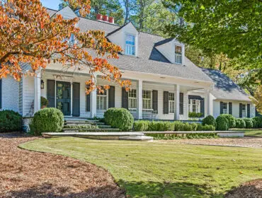 White house with dark shutters and a wraparound porch, surrounded by trees in fall foliage.