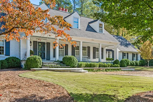 White house with dark shutters and a wraparound porch, surrounded by trees in fall foliage.