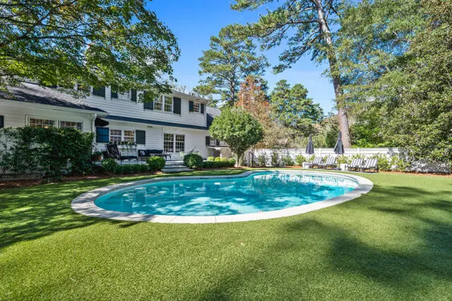 White house with green shutters and a kidney-shaped pool surrounded by manicured lawn and trees.