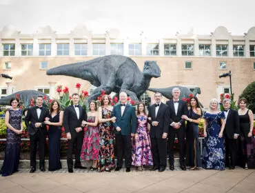 Group in formal wear poses in front of dinosaur statues and a brick building.