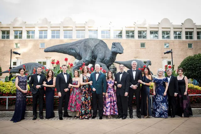 Group in formal wear poses in front of dinosaur statues and a brick building.