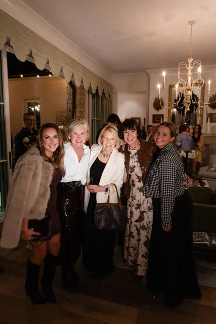 Five women posing indoors, one in a tan fur coat, another in a white shirt and sequined skirt.
