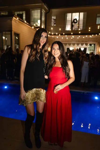 Two women posing by a pool at night. One wears a black dress with gold fringe and black boots.