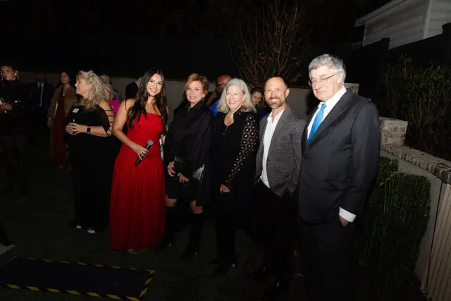 Group of people standing on grass at night, one woman in red dress holds microphone.