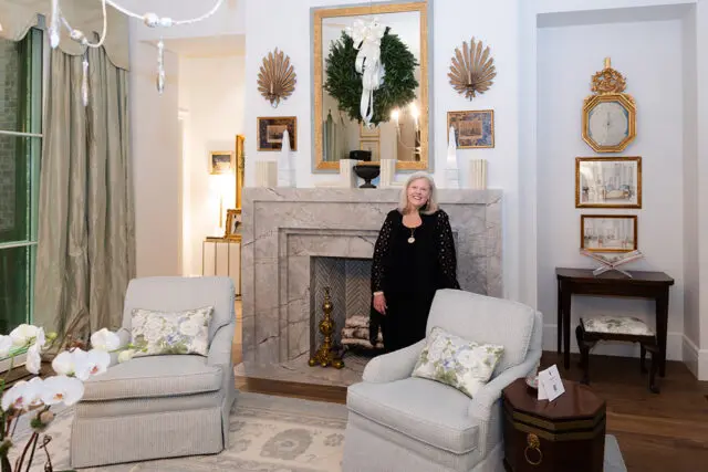 Woman in black standing by marble fireplace with wreath above.