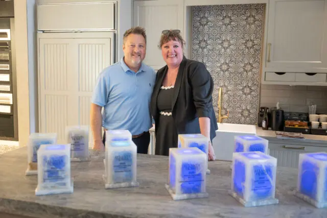 Two people stand behind a counter with awards. Kitchen cabinets and backsplash visible.