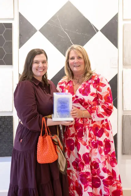 Two women holding an award in front of a black and white diamond pattern wall.