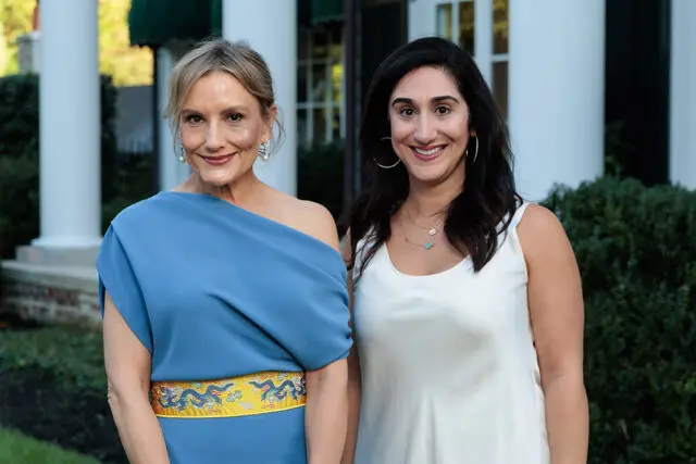 Two women smiling outdoors, one in a blue dress with a dragon belt, the other in a white dress.