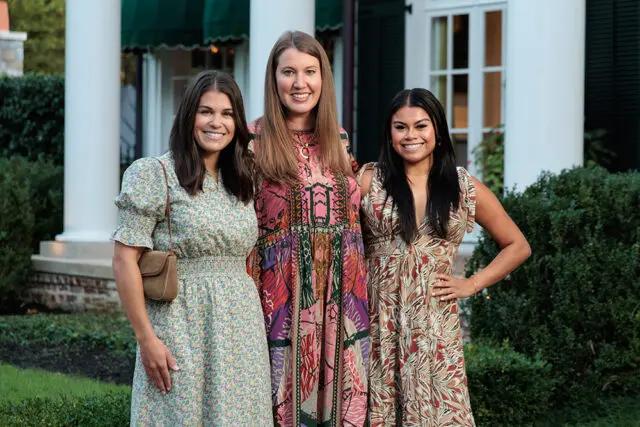 Three women smiling in front of a white house with columns and green awnings.
