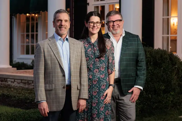 Three people stand outside a white house with columns.