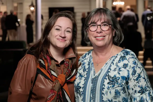Two women smiling at the camera, one in a brown leather jacket and patterned scarf, the other in a