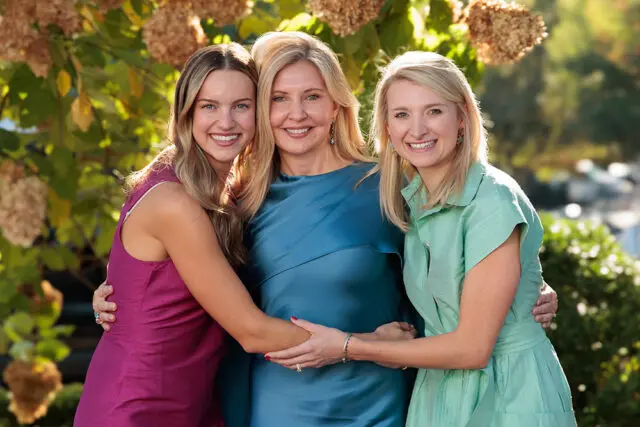 Three women smiling and embracing outdoors with foliage in the background.