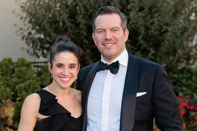 Couple in formal wear smiling outdoors with greenery in the background.