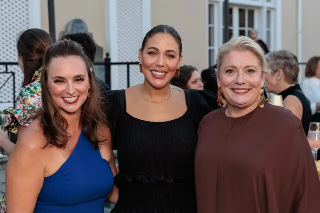 Three women smiling at an outdoor event. The woman on the left wears a blue dress. The woman in the