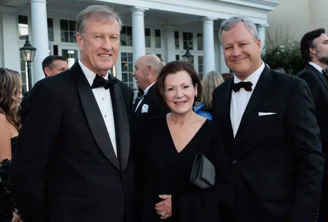 Three people in formal wear posing outdoors in front of a white building with columns.