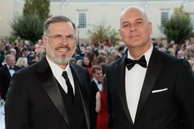 Two men in tuxedos at an event. The man on the left wears glasses and has a beard.