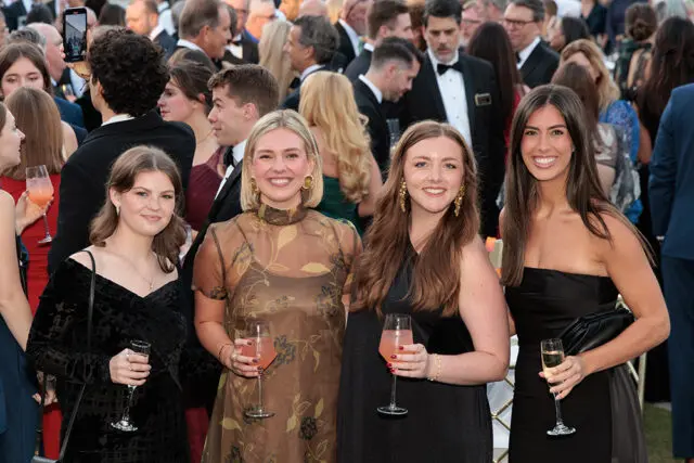 Four women in formal attire holding drinks at an outdoor event.