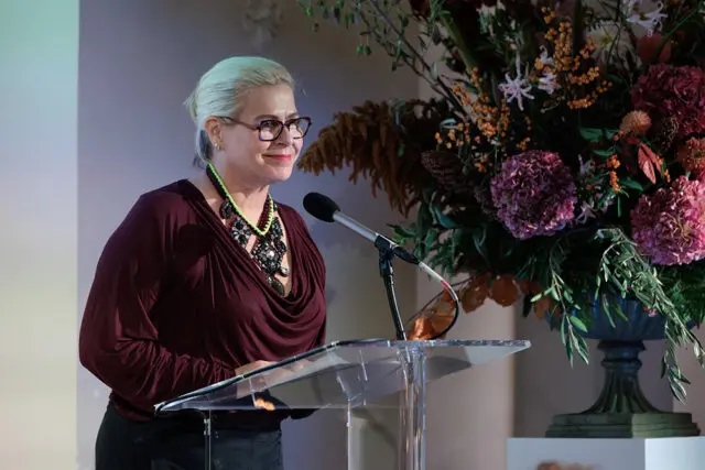 Woman with blonde hair and glasses speaking at a podium, wearing a maroon top and statement
