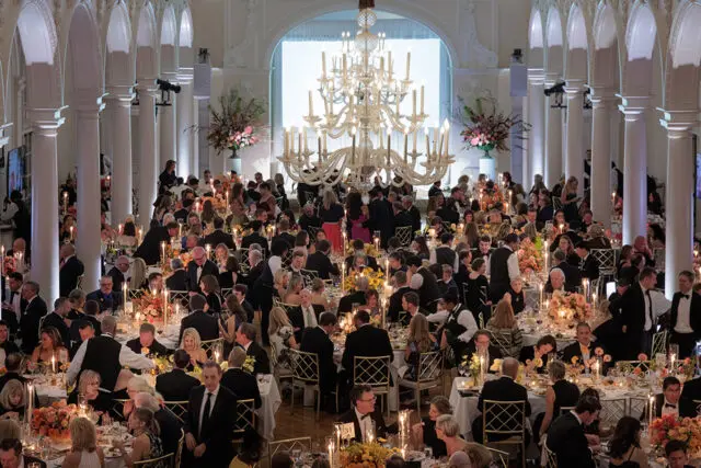 Grand ballroom with long tables set for a formal dinner, ornate chandelier, and white columns.