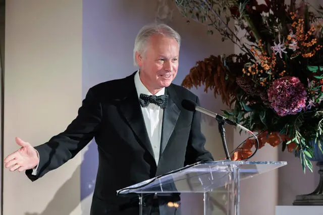 Man in tuxedo speaking at a podium with a floral arrangement
