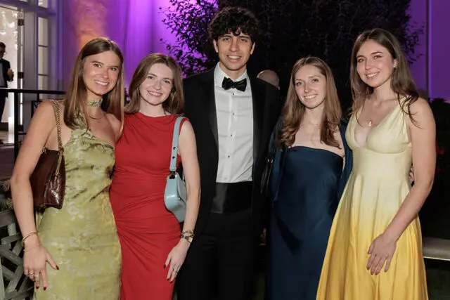 Five young adults posing for a photo. A man in a tuxedo stands between four women in formal dresses.
