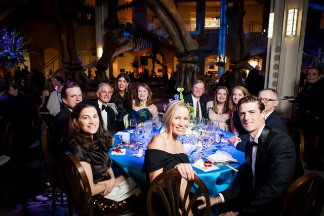 People seated at a blue-clothed table in a museum with dinosaur skeletons in the background.