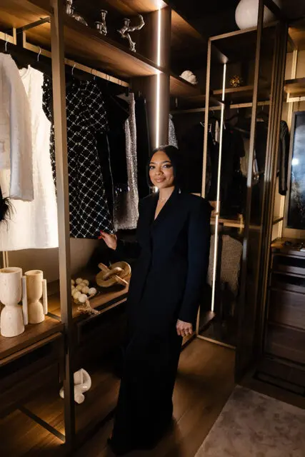 Woman in black blazer dress in walk-in closet with dark wood shelves and clothing