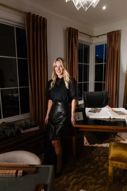 Woman in black blouse and leather skirt standing in office with dark wood desk.