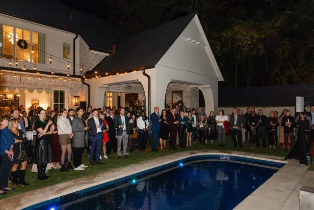 Crowd gathered by a pool at night in front of a white brick house with string lights